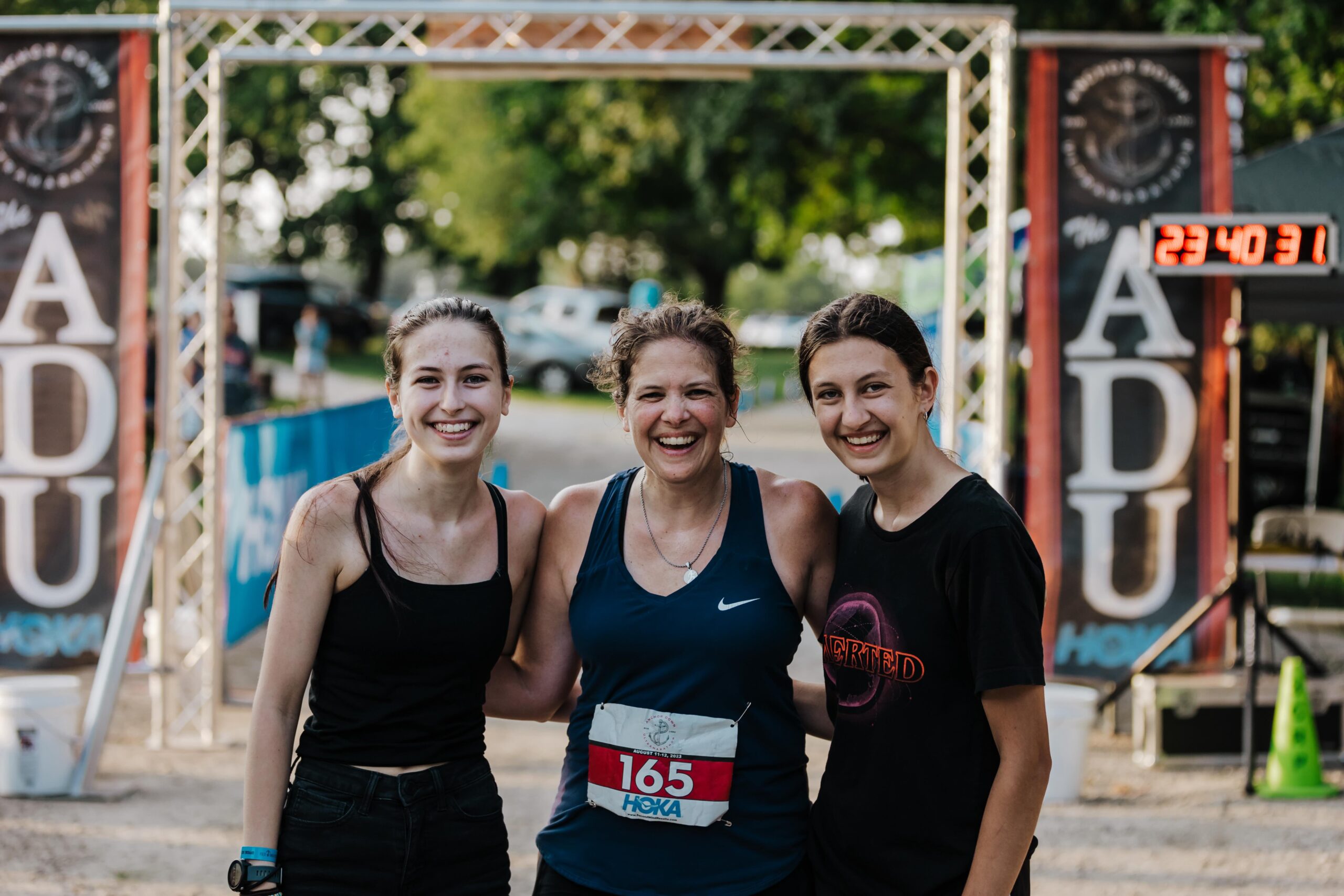 Three smiling volunteers at the Anchor Down Ultra Marathon finish line, celebrating the runners' achievements.